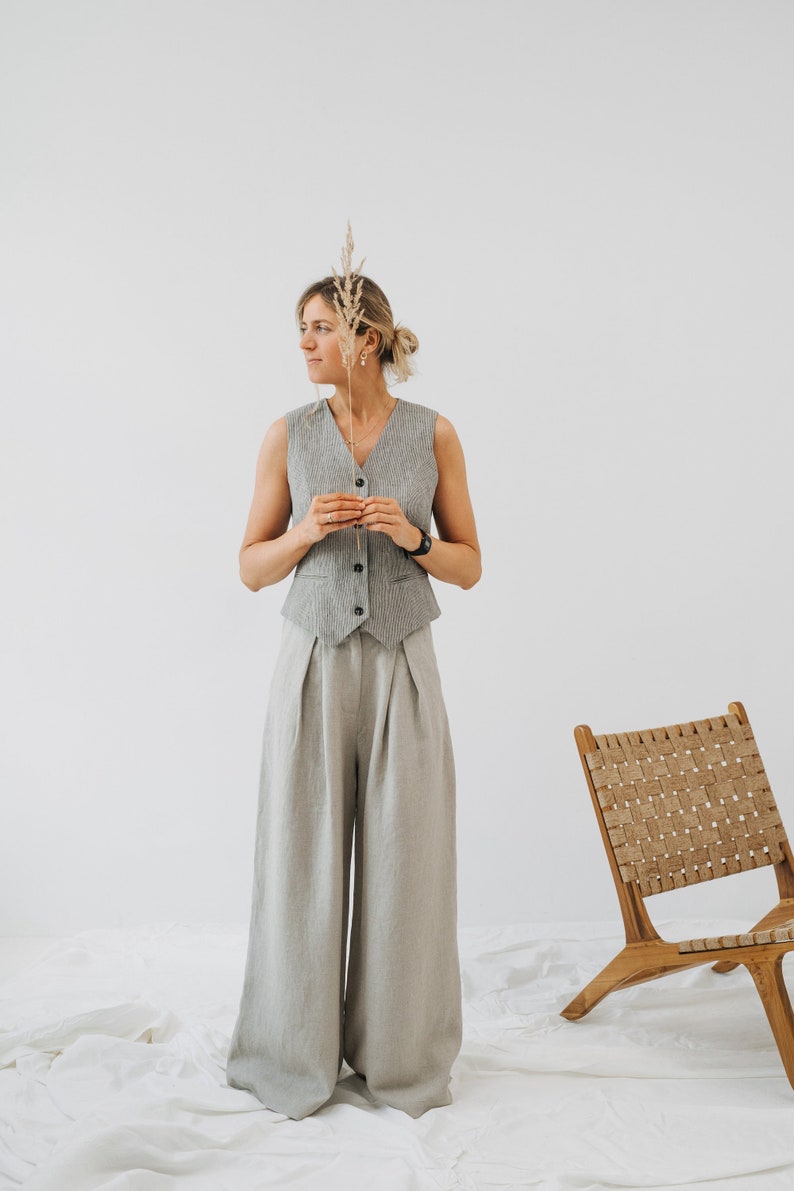 May include: A woman wearing a gray linen vest and matching wide-leg pants. She is holding a dried grass stalk in her hand and looking to the side. A wooden chair with a woven seat is in the background.