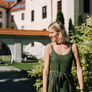 K&ouml;nnte beinhalten: Eine Frau in einem gr&uuml;nen Leinenkleid steht vor einem Geb&auml;ude mit einem roten Ziegeldach. Sie l&auml;chelt und schaut zur Seite.