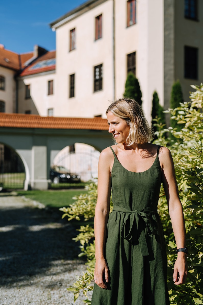 K&ouml;nnte beinhalten: Eine Frau in einem gr&uuml;nen Leinenkleid steht vor einem Geb&auml;ude mit einem roten Ziegeldach. Sie l&auml;chelt und schaut zur Seite.