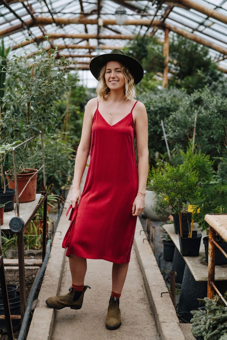 May include: A woman wearing a red slip dress and a green fedora hat stands in a greenhouse. She is wearing brown ankle boots and a gold necklace.