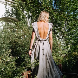 May include: A woman wearing a light gray linen dress with a low back and a bow tie. The dress has ruffles on the shoulders and a flowing skirt. She is standing in a garden with green foliage and a wooden bench.