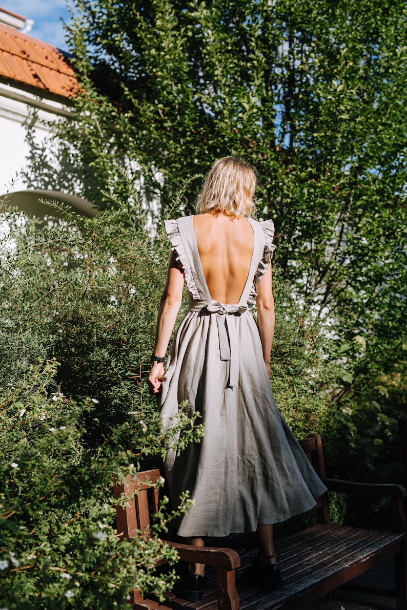 May include: A woman wearing a light gray linen dress with a low back and a bow tie. The dress has ruffles on the shoulders and a flowing skirt. She is standing in a garden with green foliage and a wooden bench.