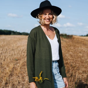 May include: A woman wearing a black hat, a green cardigan, a white tank top, and denim shorts stands in a field of dry grass. She is smiling and has a small bouquet of yellow flowers tucked into the pocket of her cardigan.