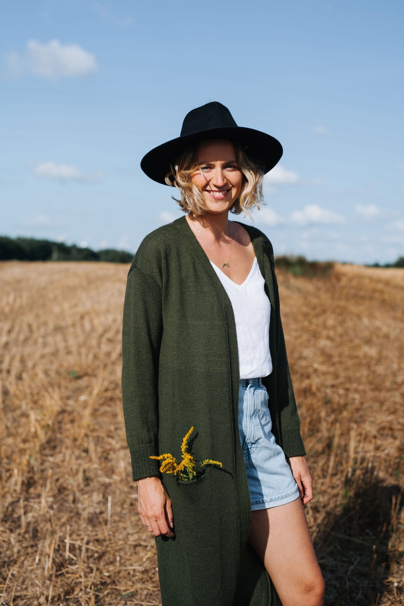 May include: A woman wearing a black hat, a green cardigan, a white tank top, and denim shorts stands in a field of dry grass. She is smiling and has a small bouquet of yellow flowers tucked into the pocket of her cardigan.