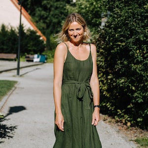 K&ouml;nnte beinhalten: Eine Frau in einem gr&uuml;nen Leinenkleid mit einem Bindeg&uuml;rtel steht auf einem Weg vor einer gr&uuml;nen Hecke.