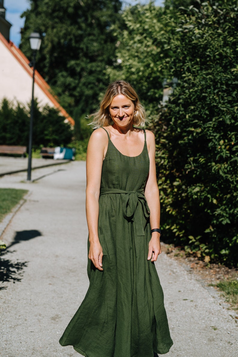K&ouml;nnte beinhalten: Eine Frau in einem gr&uuml;nen Leinenkleid mit einem Bindeg&uuml;rtel steht auf einem Weg vor einer gr&uuml;nen Hecke.