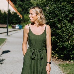 K&ouml;nnte beinhalten: Eine Frau in einem gr&uuml;nen Leinenkleid mit einem Bindeg&uuml;rtel. Sie steht auf einem Weg mit gr&uuml;nen B&auml;umen im Hintergrund.