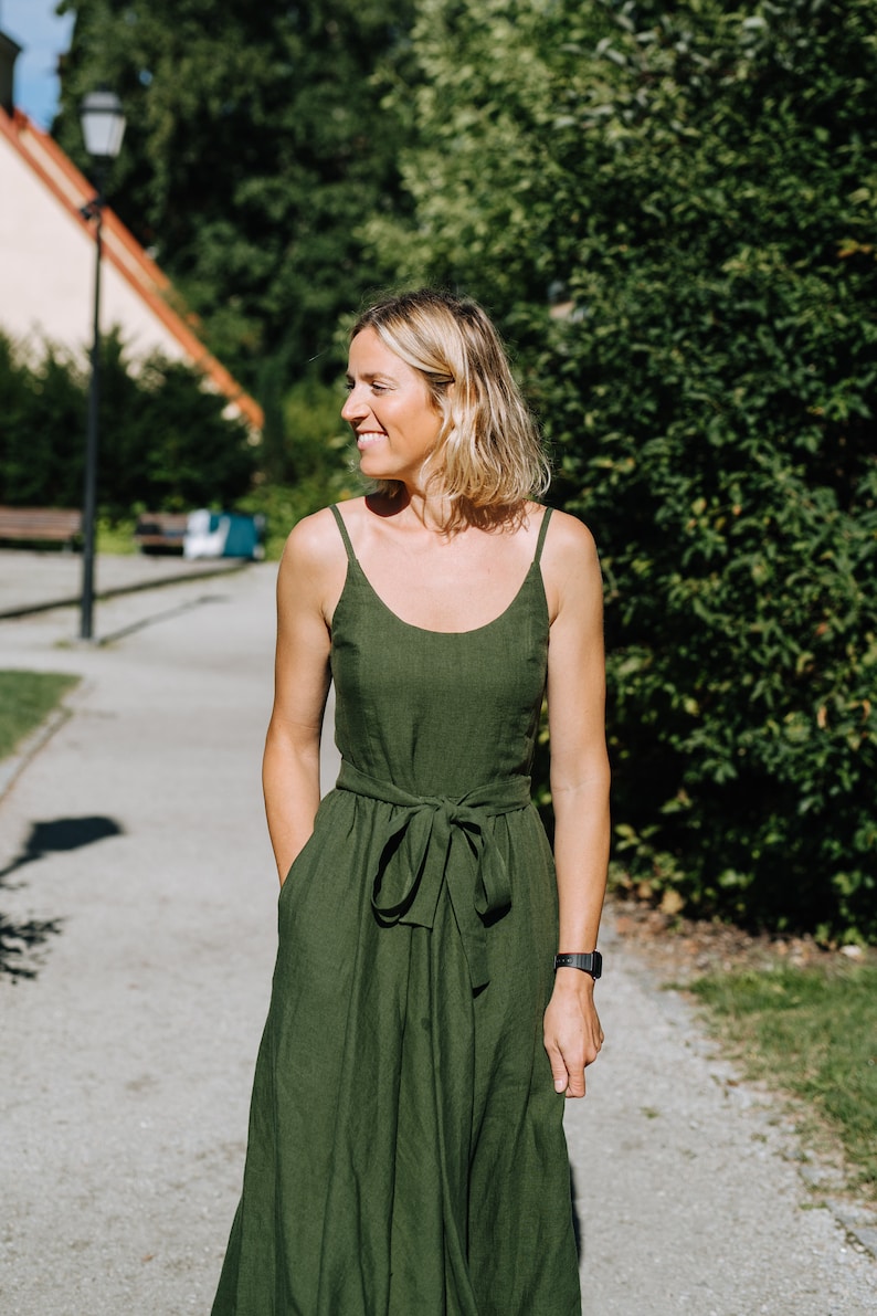 K&ouml;nnte beinhalten: Eine Frau in einem gr&uuml;nen Leinenkleid mit einem Bindeg&uuml;rtel. Sie steht auf einem Weg mit gr&uuml;nen B&auml;umen im Hintergrund.