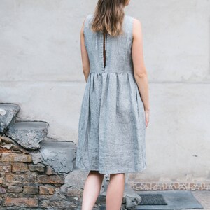 May include: A woman wearing a gray linen dress with a back slit and a gathered waist. She is wearing brown sandals and is standing on a brick walkway.