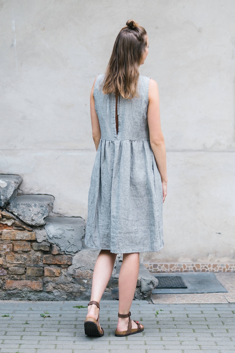 May include: A woman wearing a gray linen dress with a back slit and a gathered waist. She is wearing brown sandals and is standing on a brick walkway.