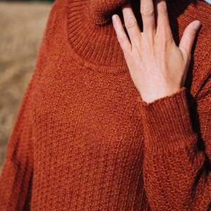May include: A close-up of a rust-coloured turtleneck sweater with a ribbed knit texture. The sweater is made of a soft, warm material and has a relaxed fit.