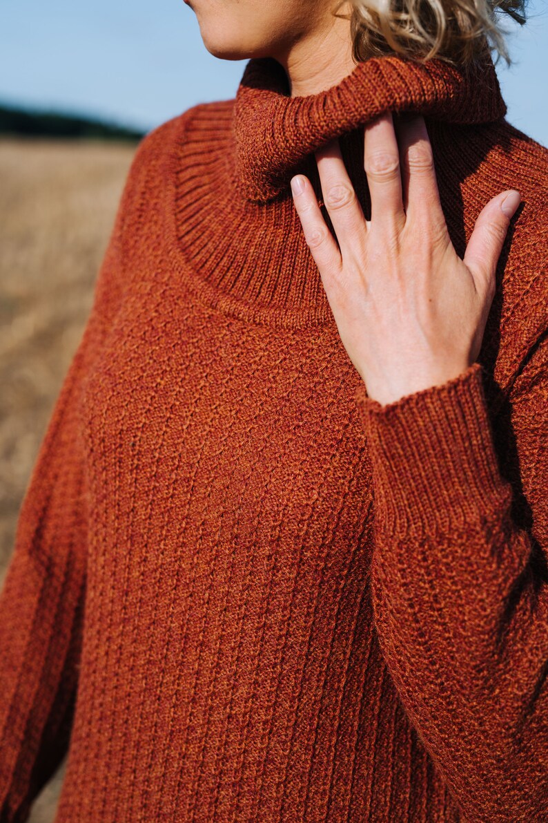 May include: A close-up of a rust-coloured turtleneck sweater with a ribbed knit texture. The sweater is made of a soft, warm material and has a relaxed fit.
