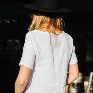 Op de afbeelding: Een persoon draagt een wit linnen shirt, een zwarte jeans en een zwarte fedora.
