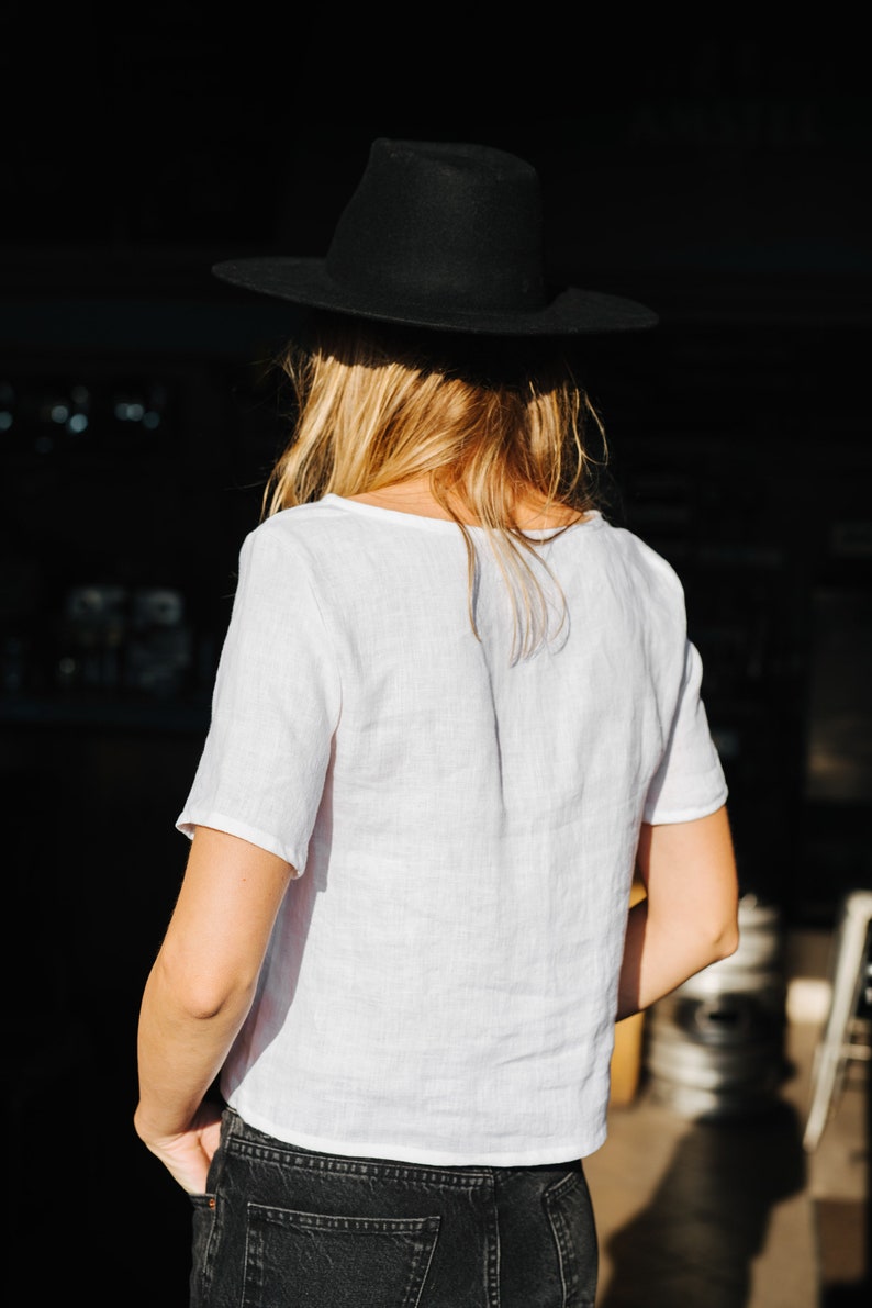 Op de afbeelding: Een persoon draagt een wit linnen shirt, een zwarte jeans en een zwarte fedora.