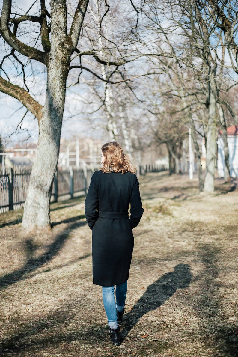 May include: A woman wearing a black coat and blue jeans walks through a park with bare trees.