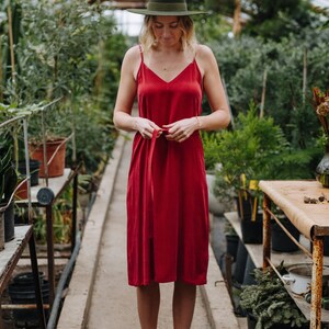 May include: A woman wearing a red dress with a green hat and brown boots stands in a greenhouse. The dress has a thin red ribbon tied at the waist.