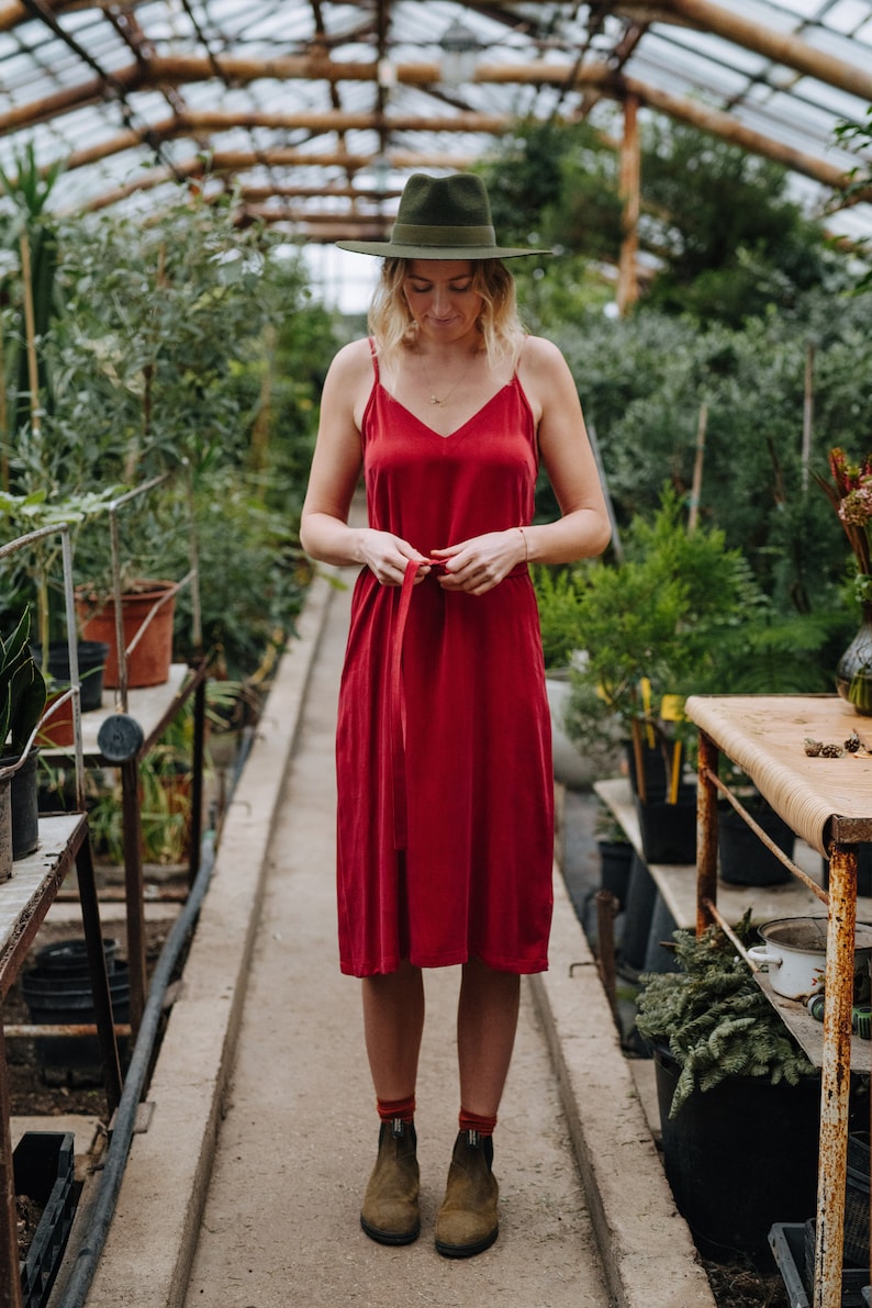 May include: A woman wearing a red dress with a green hat and brown boots stands in a greenhouse. The dress has a thin red ribbon tied at the waist.