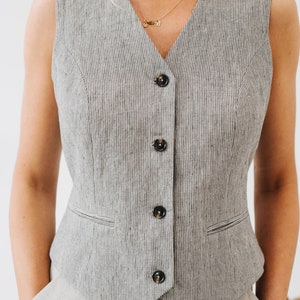 May include: A sleeveless vest with a V-neckline and a button-up front. The vest is made of a striped fabric in a light gray and black color.