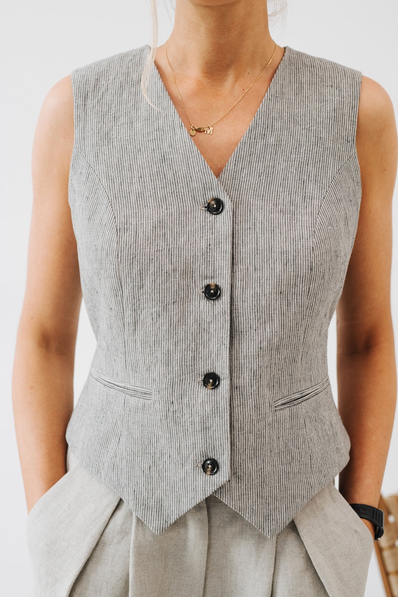 May include: A sleeveless vest with a V-neckline and a button-up front. The vest is made of a striped fabric in a light gray and black color.