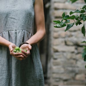 May include: A person wearing a gray linen dress holds a handful of small green fruits in their hands.