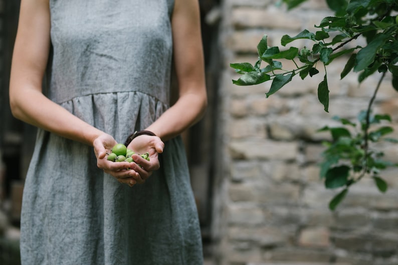 May include: A person wearing a gray linen dress holds a handful of small green fruits in their hands.