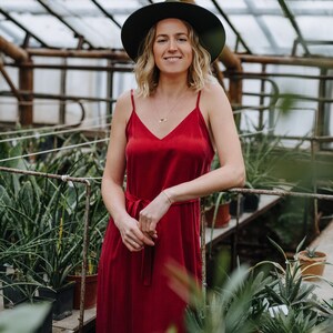 May include: A woman wearing a red dress and a green hat stands in a greenhouse. She is surrounded by potted plants.