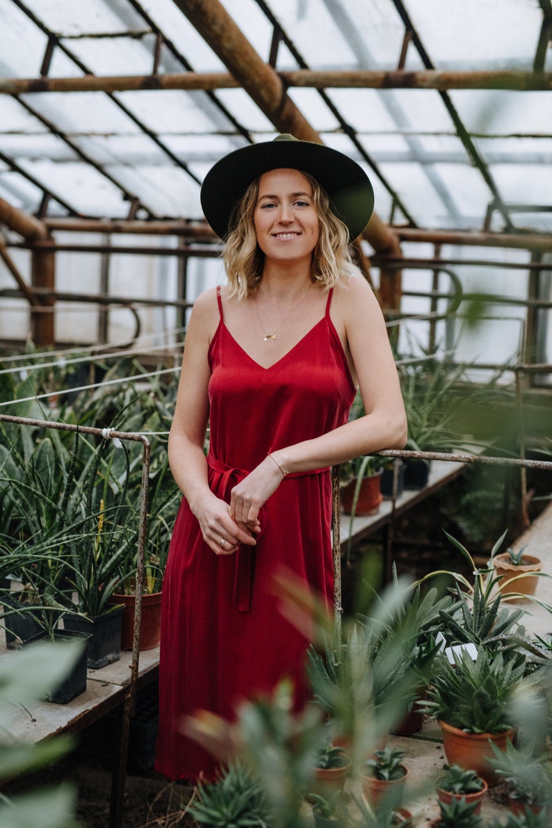 May include: A woman wearing a red dress and a green hat stands in a greenhouse. She is surrounded by potted plants.