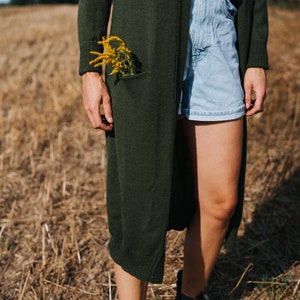 May include: A person wearing a long, dark green cardigan with a bouquet of yellow flowers tucked into the pocket. They are also wearing light blue denim shorts and brown leather ankle boots.