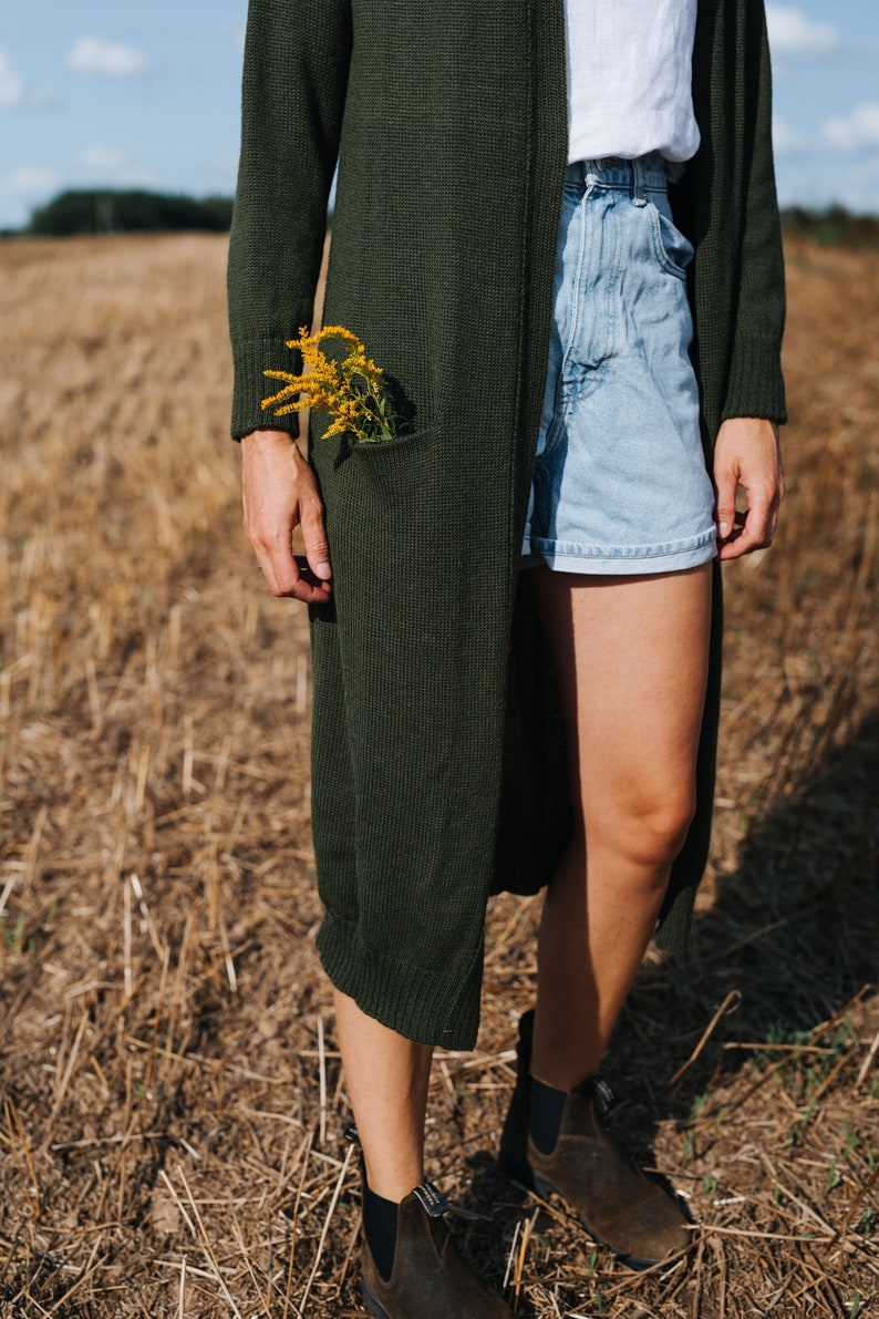 May include: A person wearing a long, dark green cardigan with a bouquet of yellow flowers tucked into the pocket. They are also wearing light blue denim shorts and brown leather ankle boots.