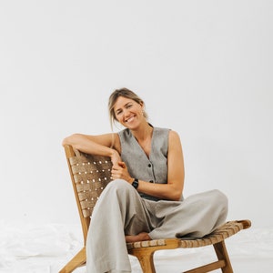 May include: A woman sits in a woven wood chair with a light gray sleeveless vest and matching wide-leg pants. She is smiling at the camera.
