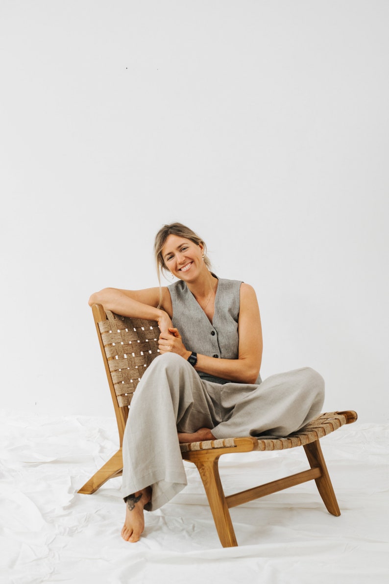 May include: A woman sits in a woven wood chair with a light gray sleeveless vest and matching wide-leg pants. She is smiling at the camera.