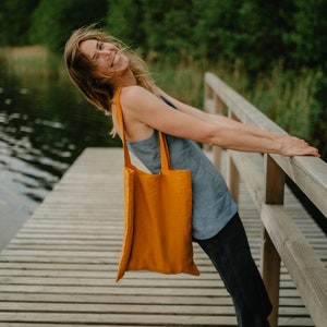 May include: A woman wearing a blue tank top and black pants is standing on a wooden dock. She is holding a yellow tote bag and smiling.