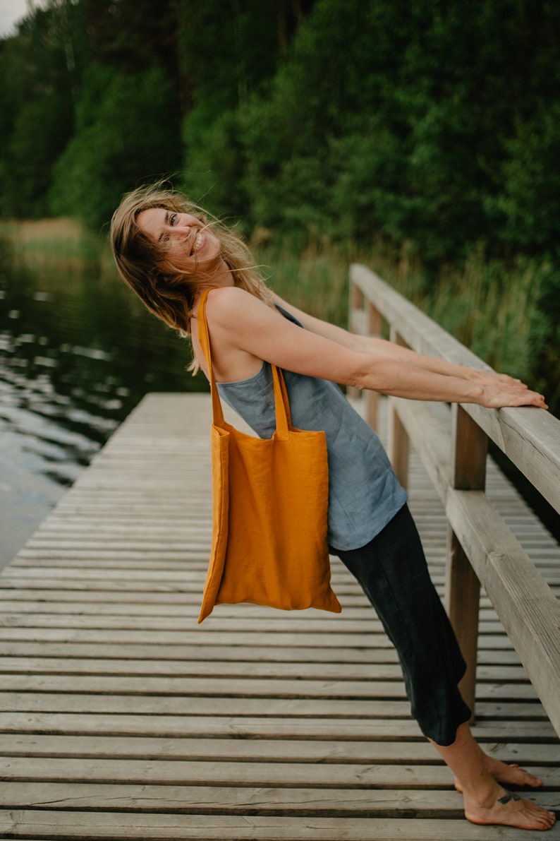 May include: A woman wearing a blue tank top and black pants is standing on a wooden dock. She is holding a yellow tote bag and smiling.