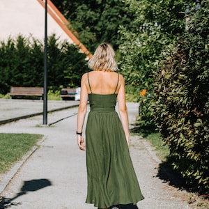 K&ouml;nnte beinhalten: Eine Frau in einem gr&uuml;nen Maxikleid geht auf einem gepflasterten Weg in einem Park entlang. Der Weg ist von gr&uuml;nen B&uuml;schen und B&auml;umen ges&auml;umt.