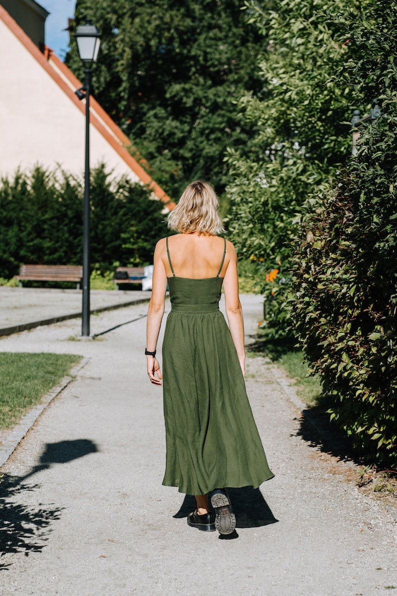 K&ouml;nnte beinhalten: Eine Frau in einem gr&uuml;nen Maxikleid geht auf einem gepflasterten Weg in einem Park entlang. Der Weg ist von gr&uuml;nen B&uuml;schen und B&auml;umen ges&auml;umt.