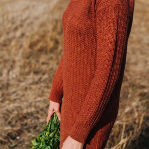 May include: A rust-coloured turtleneck sweater dress with a ribbed texture. The dress is being worn by a person holding a bouquet of green flowers.