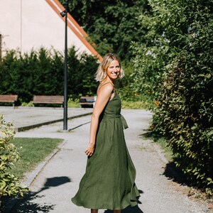 K&ouml;nnte beinhalten: Eine Frau in einem gr&uuml;nen Leinenkleid geht auf einem Weg in einem Park spazieren. Sie l&auml;chelt und schaut &uuml;ber ihre Schulter.