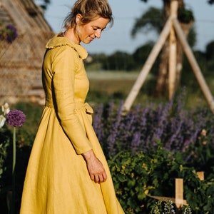 Pode incluir: Uma mulher com um vestido de linho amarelo caminha por um jardim. O vestido tem um colarinho com babados e uma cintura cintada. O jardim est&aacute; cheio de flores roxas e plantas verdes.
