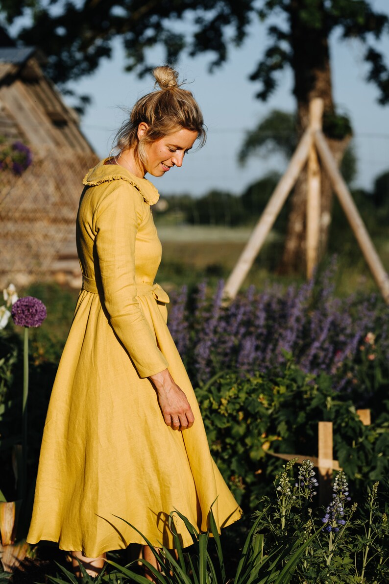 Pode incluir: Uma mulher com um vestido de linho amarelo caminha por um jardim. O vestido tem um colarinho com babados e uma cintura cintada. O jardim est&aacute; cheio de flores roxas e plantas verdes.