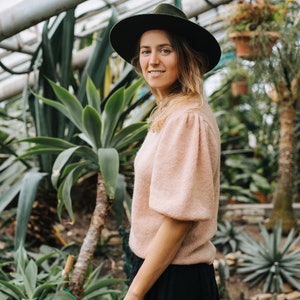 May include: A woman wearing a pink sweater and a black skirt stands in a greenhouse. She is wearing a wide-brimmed black hat and looking at the camera. The background is filled with lush green plants.