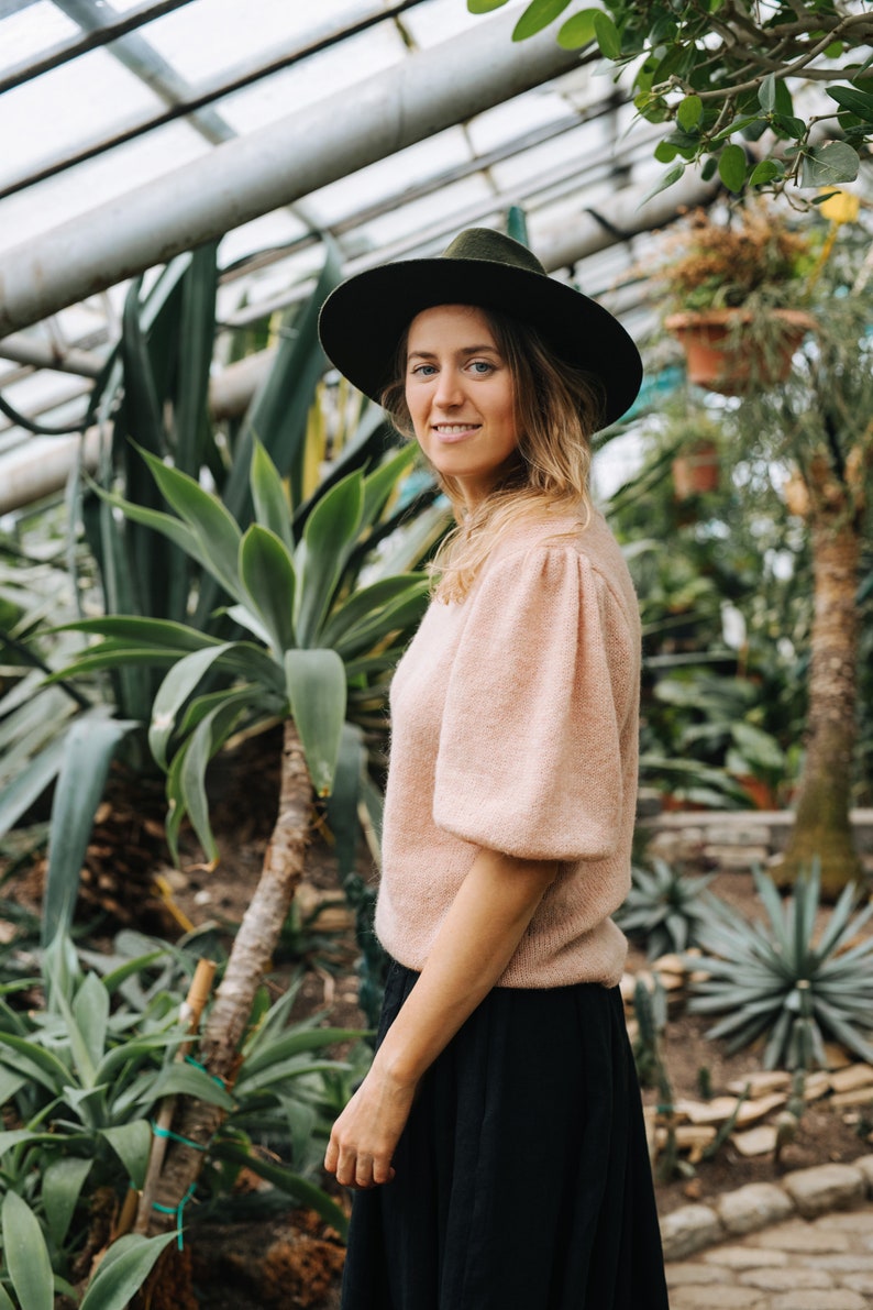 May include: A woman wearing a pink sweater and a black skirt stands in a greenhouse. She is wearing a wide-brimmed black hat and looking at the camera. The background is filled with lush green plants.