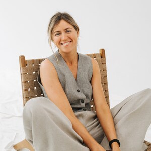 May include: A woman wearing a gray sleeveless vest and light gray pants sits in a woven wooden chair. She is smiling and looking at the camera.