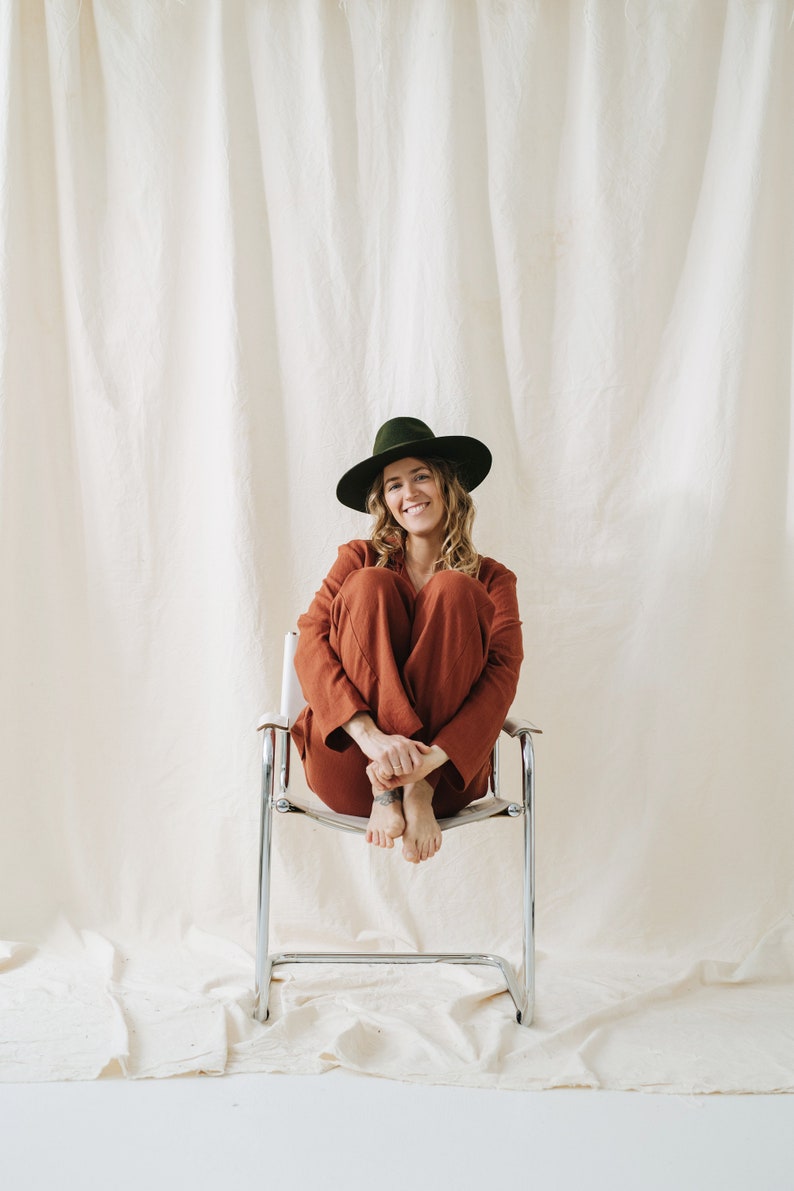 Puede incluir: Una mujer con un mono de color &oacute;xido y un sombrero fedora verde oscuro se sienta en una silla plateada con las piernas cruzadas. Sonr&iacute;e a la c&aacute;mara.