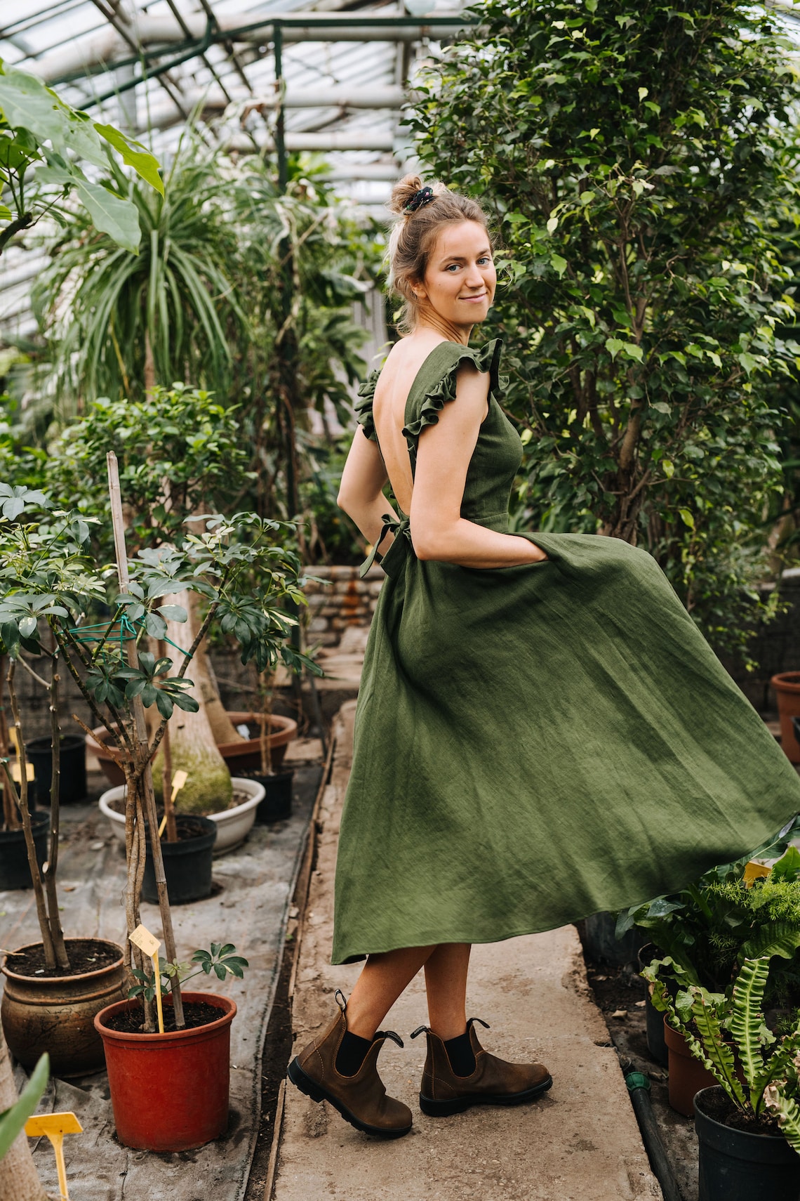 May include: A woman in a green linen dress stands in a greenhouse. She is wearing brown ankle boots and has her hair pulled back in a bun. The dress has a low back and a ruffled neckline.