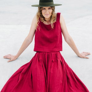 May include: A woman wearing a red linen top and skirt with a wide-brimmed green hat. She is sitting on a white surface.