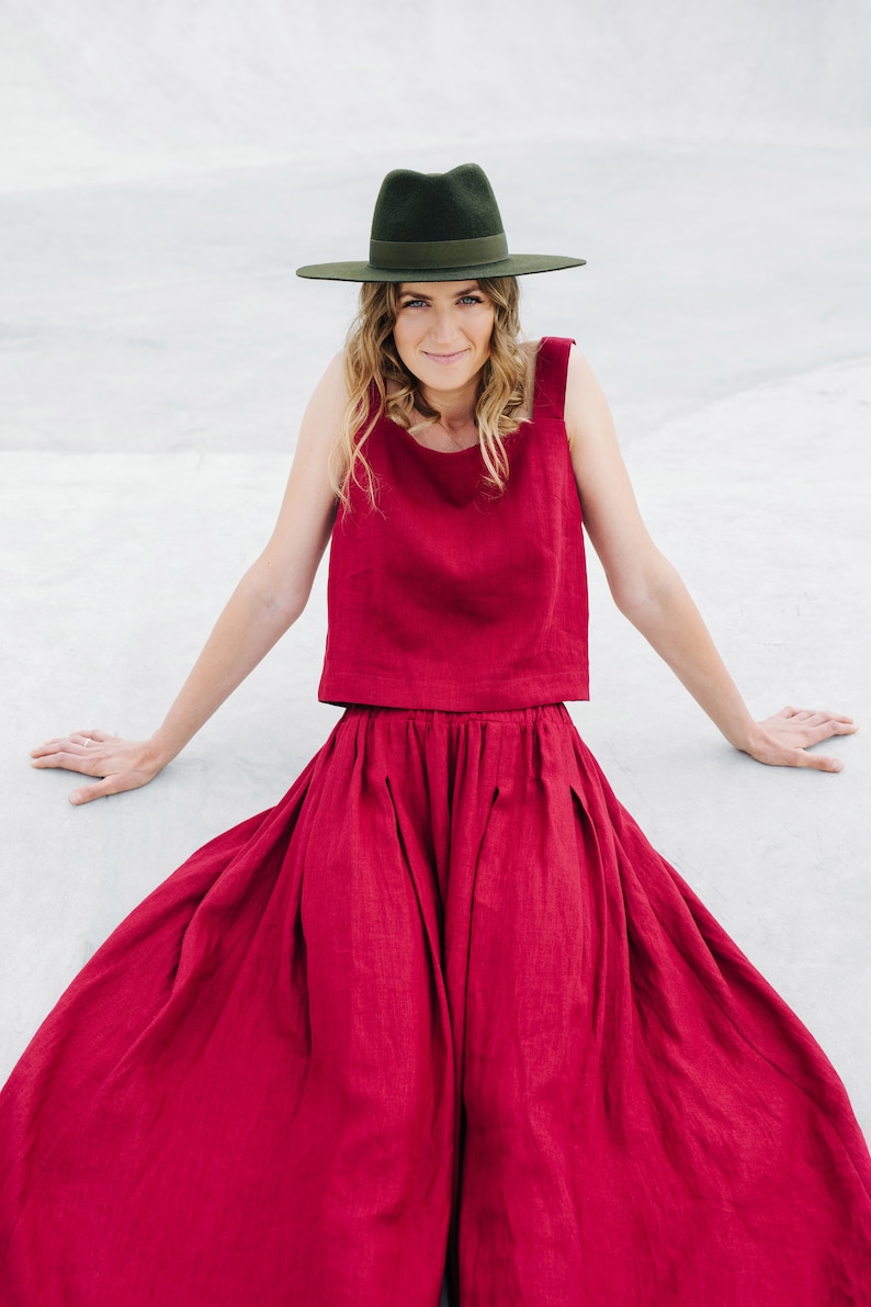 May include: A woman wearing a red linen top and skirt with a wide-brimmed green hat. She is sitting on a white surface.