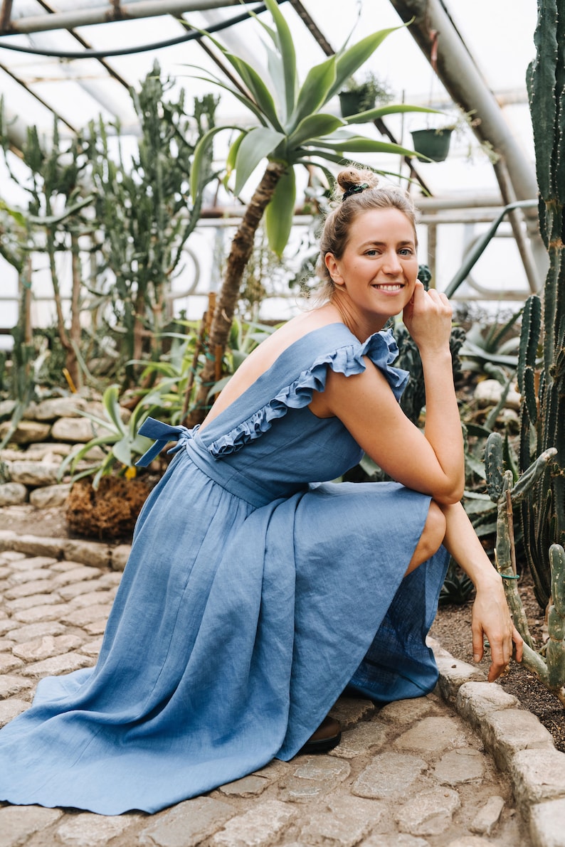 Puede incluir: Una mujer con un vestido de lino azul con volantes posa en un invernadero. El vestido tiene la espalda baja y un lazo en la cintura. Est&aacute; rodeada de varias plantas, incluyendo cactus.
