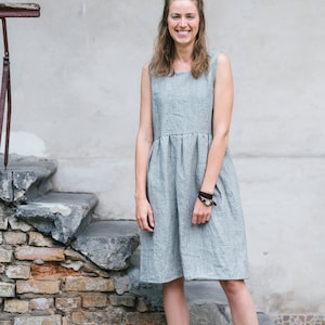 May include: A woman wearing a gray linen dress and brown sandals stands in front of a stone wall. She is smiling and looking at the camera.