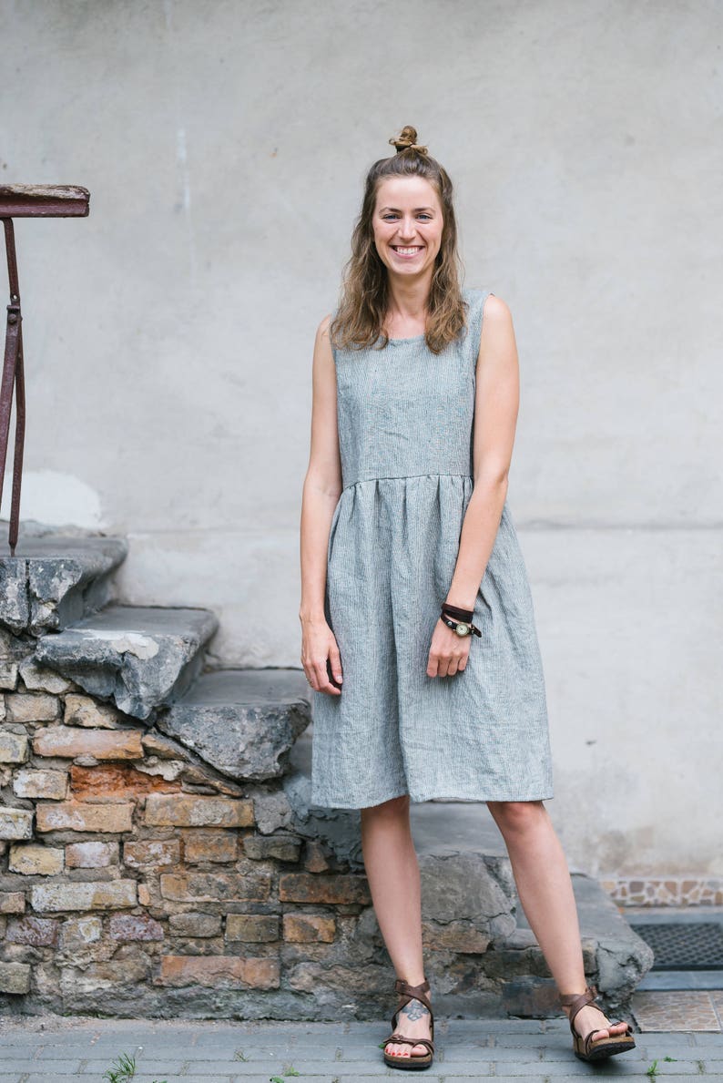 May include: A woman wearing a gray linen dress and brown sandals stands in front of a stone wall. She is smiling and looking at the camera.