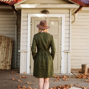 May include: A woman wearing a green linen dress and a brown hat stands in front of a white wooden building with a red roof.  She is walking on a wooden deck covered in fallen autumn leaves.